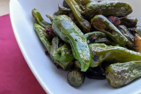 A pile of glistening, fried padron peppers in a white bowl, some with slightly charred edges, hinting at a delicious, savory snack.