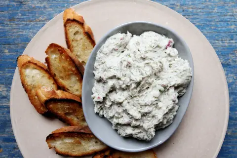 A bowl of creamy bluefish pate with herbs and red onion, served with toasted baguette slices on a light-colored plate.