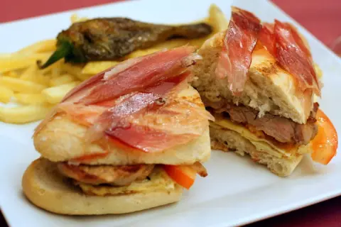 A delicious bocadillo with tender meat, cured ham, and fresh tomato, served with crispy fries and a roasted pepper.