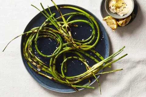 Grilled garlic scapes, seasoned with sea salt, are artfully arranged on a dark plate, with a small bowl of creamy dip and lemon wedges nearby.