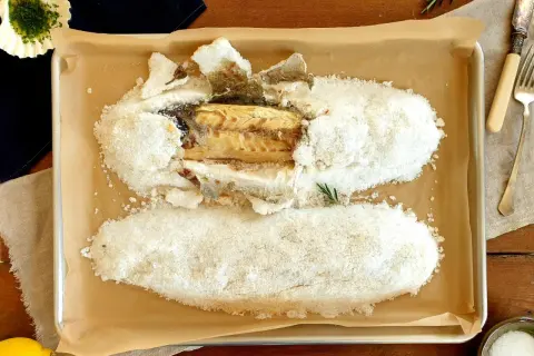 A whole salt crusted sea bass, baked to perfection, rests on a baking sheet. A lemon and herbs complete the dish.