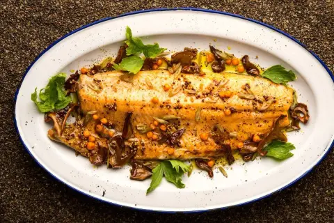 A beautifully broiled trout, glistening with olive oil and garnished with mustard seeds, sea buckthorn berries, and fresh herbs on a white enamel platter.