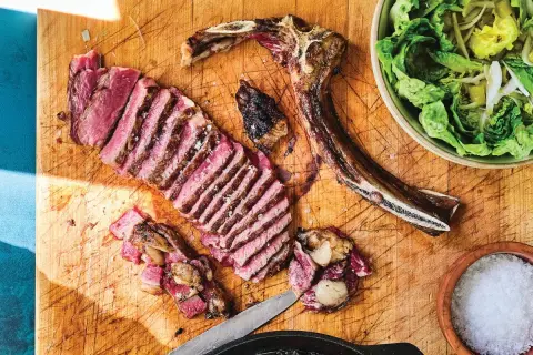 A perfectly cooked Spanish steak, sliced and ready to enjoy, sits on a wooden board with a side of fresh salad and a bowl of coarse salt.