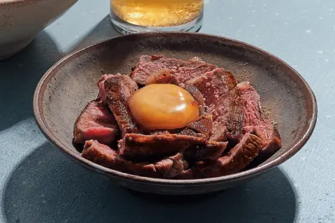 Sliced steak in a bowl topped with a glistening salt cured egg yolk, ready to be mixed into a rich, savory dish.