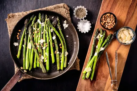 A pan of cooked asparagus with shaved parmesan and pine nuts, showing how to make asparagus a delicious side dish.