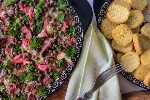 Thinly sliced beef carpaccio, adorned with capers and parsley, is served with toasted bread rounds.