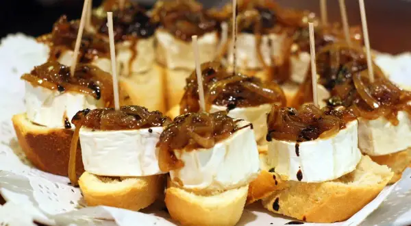 Delicious pincho recipes: bite-sized bread topped with creamy cheese, caramelized onions, and a balsamic glaze, all secured with a toothpick.