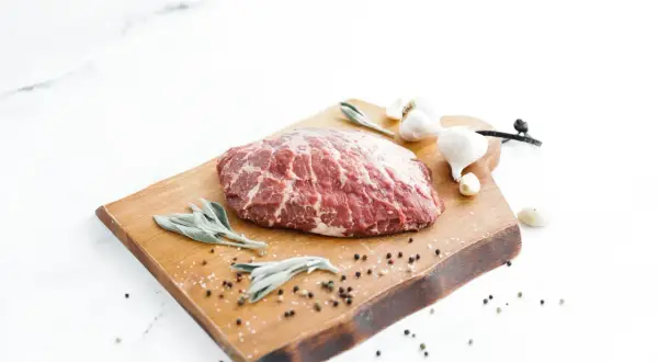 A beautiful cut of presa ibérica sits on a wooden board with sage leaves, garlic, and peppercorns, ready for a delicious meal.