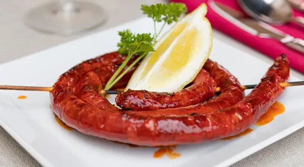 A coiled chistorra sausage, grilled to perfection, served on a white plate with a lemon wedge and parsley. A delicious chistorra recipe.