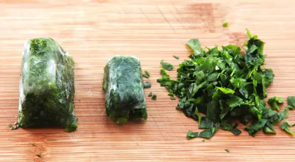 Learn how to freeze fresh herbs: two frozen herb blocks and a pile of chopped herbs on a wooden cutting board.