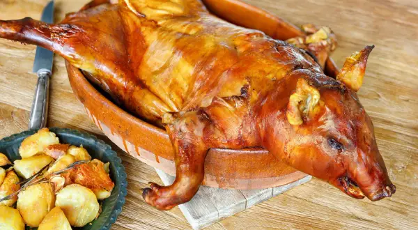 A whole roasted suckling pig, golden brown and glistening, sits in a rustic clay dish. This image evokes delicious suckling pig recipes and a feast.
