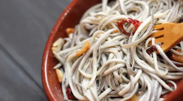A close-up of a rustic bowl filled with a tangle of little eel, glistening with garlic and chili flakes. A wooden fork is nestled within the dish.