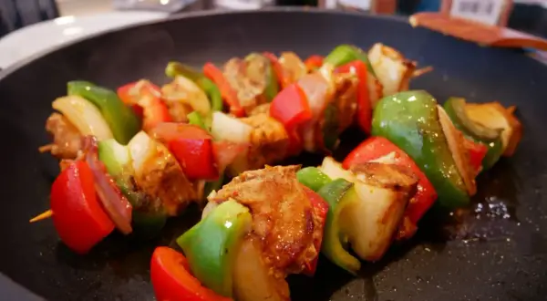 Pinchos morunos sizzling in a pan, with chunks of marinated meat, red and green bell peppers, and onions on wooden skewers.