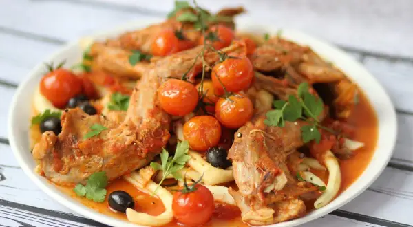 A hearty rabbit stew with tender meat, cherry tomatoes, olives, and pasta, garnished with fresh parsley.