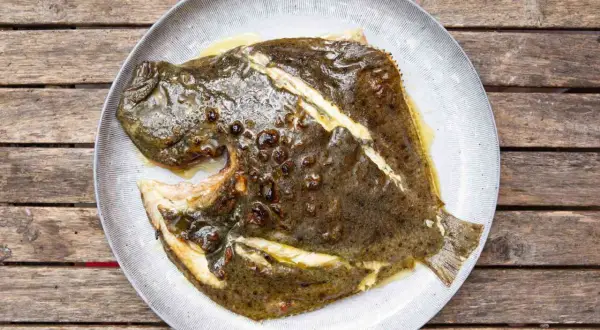 A whole grilled turbot, glistening with butter and herbs, served on a rustic plate atop a weathered wooden table.