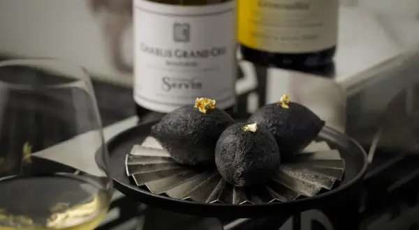 A glass of chablis white wine sits beside a plate of three dark, spherical treats topped with gold leaf, with bottles of chablis grand cru in the background.