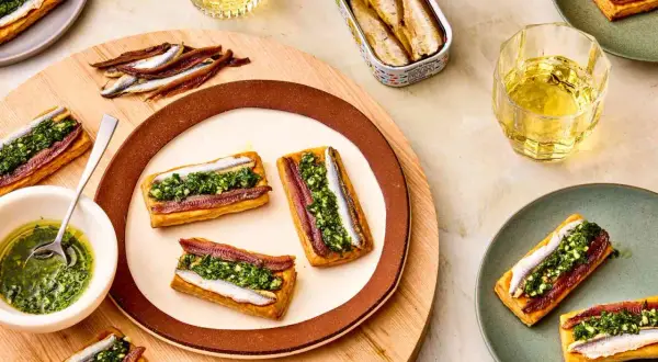 Rectangular pastries topped with red and white anchovies and green sauce. A tin of white anchovies sits nearby.