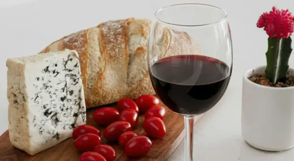 A glass of red wine, perhaps a bold trebbiano wine, sits beside a rustic loaf of bread, blue cheese, and cherry tomatoes, with a small potted cactus nearby.