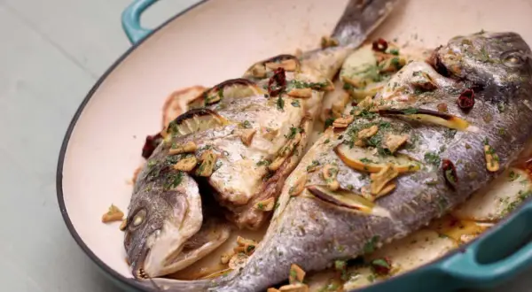 Two whole sea bream baked with lemon slices, garlic, and herbs in a teal pan, served over sliced potatoes.