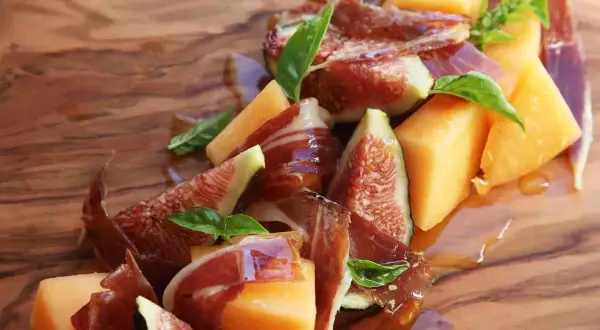 A refreshing melon con jamon appetizer with fresh figs and basil, drizzled with honey on a wooden board.