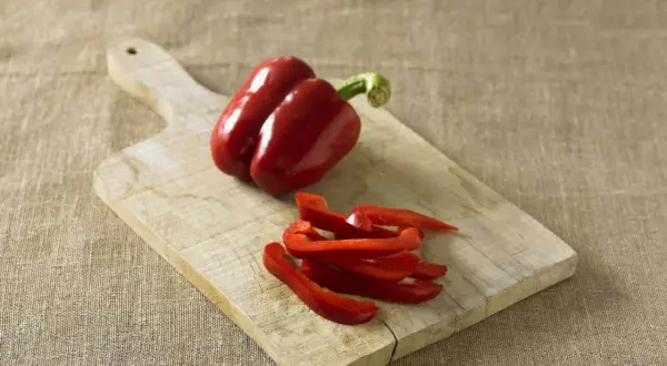 Red bell peppers, one whole and some sliced, on a wooden cutting board. Perfect for learning how to preserve peppers for later use.