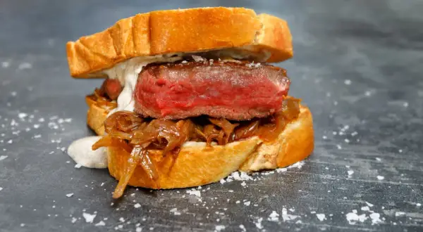 A juicy pepito de ternera, a tender steak sandwich on toasted bread with caramelized onions and a creamy sauce, sprinkled with sea salt.