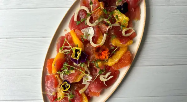 A vibrant tuna crudo dish featuring thinly sliced raw tuna, citrus segments, fennel, microgreens, and edible flowers.