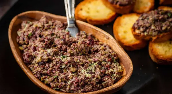A rustic wooden bowl overflows with chunky tapenade, a savory spread of olives and herbs, ready to be enjoyed with toasted crostini.