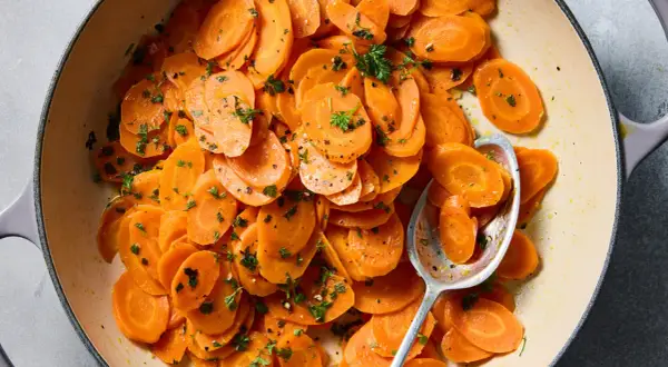 A close-up of a pot of glazed vichy carrots, glistening with butter and sprinkled with fresh parsley. A spoon rests in the pot, ready to serve.