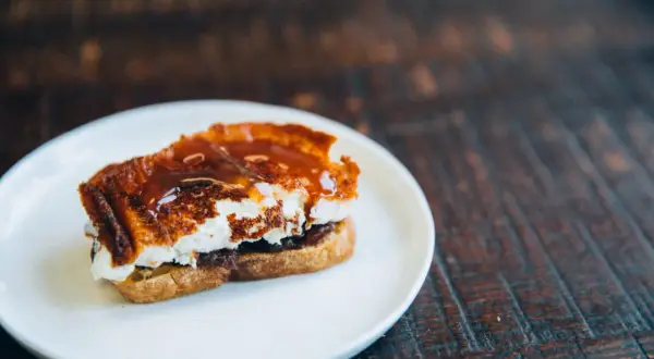 A decadent goat cheese toast, caramelized and drizzled with a rich, glossy sauce, sits on a white plate.