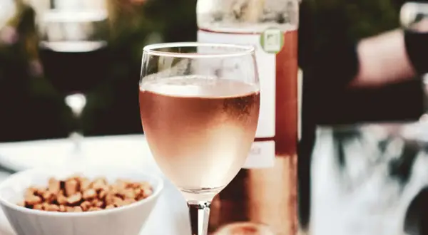 A glass of rosé wine, perhaps a Marsanne blend, sits on a table with a bowl of nuts and a bottle of wine.