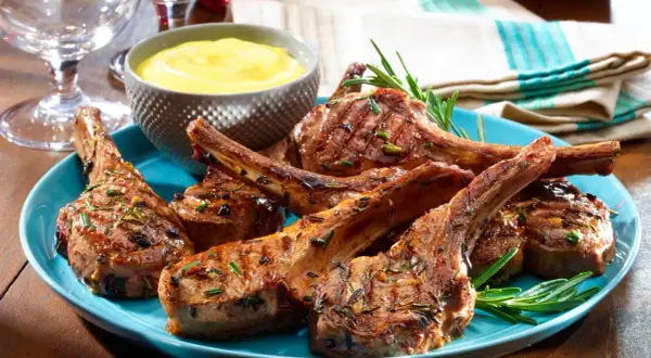 Grilled lamb chops, seasoned with rosemary, served on a blue plate with a side of yellow sauce. A taste of Spanish lamb meat.