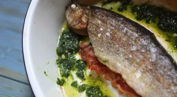 A whole la trucha, seasoned with salt, rests in a white dish with a vibrant green herb sauce and crispy bacon.