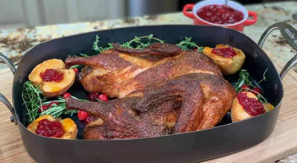 A beautifully roasted smoked duck rests in a dark roasting pan, garnished with fresh rosemary, cranberries, and baked apple halves topped with cranberry sauce.