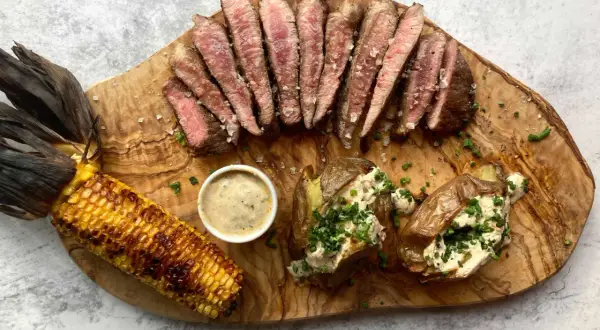 Juicy pork presa, grilled corn, and loaded baked potatoes served on a wooden board.