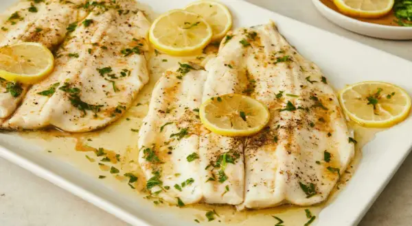 Two beautifully cooked truchas fish fillets, glistening with butter and herbs, are garnished with fresh lemon slices on a white platter.