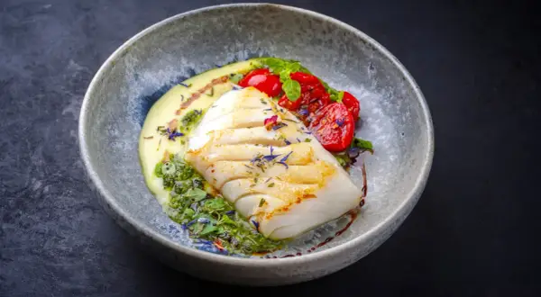 A beautifully plated poached fish recipe, featuring a white fish fillet with grilled marks, served with creamy sauce, pesto, and cherry tomatoes.