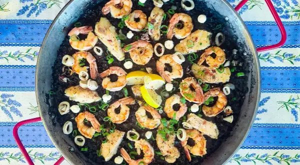 A delicious squid ink rice paella, brimming with plump shrimp, tender calamari rings, and flaky white fish, all garnished with fresh green onions and lemon wedges.