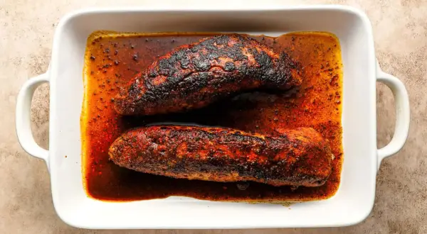 Two seasoned pork tenderloins, ready for a delicious solomillo recipe, rest in a white baking dish filled with rich, spiced juices.
