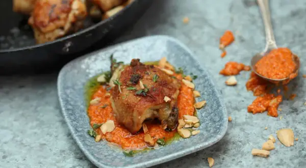 A succulent piece of chicken, part of a delicious chicken tapas dish, rests on a bed of vibrant orange sauce, garnished with herbs and chopped nuts.