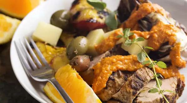 Slices of grilled spanish pork tenderloin with olives, orange wedges, and a vibrant romesco sauce.