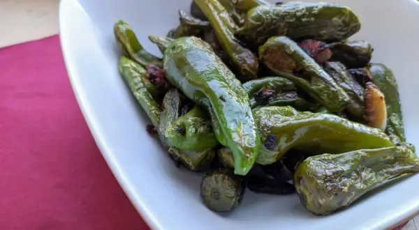 A pile of glistening, fried padron peppers in a white bowl, some with slightly charred edges, hinting at a delicious, savory snack.