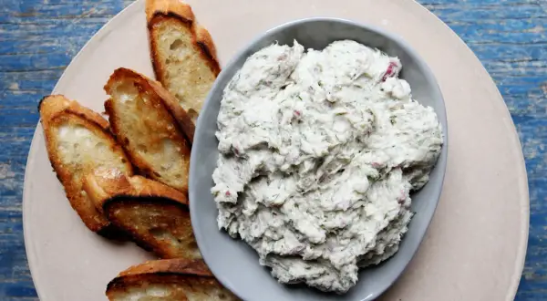 A bowl of creamy bluefish pate with herbs and red onion, served with toasted baguette slices on a light-colored plate.