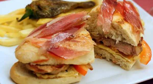 A delicious bocadillo with tender meat, cured ham, and fresh tomato, served with crispy fries and a roasted pepper.