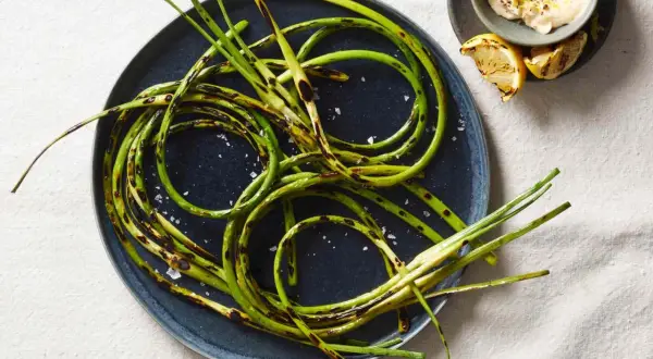 Grilled garlic scapes, seasoned with sea salt, are artfully arranged on a dark plate, with a small bowl of creamy dip and lemon wedges nearby.