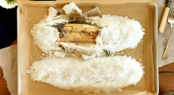 A whole salt crusted sea bass, baked to perfection, rests on a baking sheet. A lemon and herbs complete the dish.