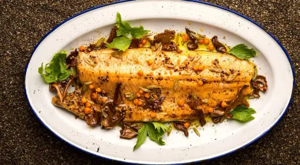 A beautifully broiled trout, glistening with olive oil and garnished with mustard seeds, sea buckthorn berries, and fresh herbs on a white enamel platter.