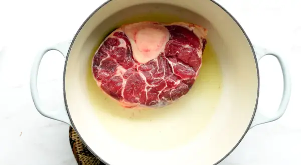 A thick beef shank, or osso buco in Spanish, is browning in a light-colored pot with oil.