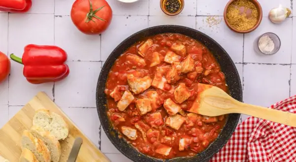 Tuna in tomato sauce simmering in a pan, with fresh tomatoes, red bell peppers, and crusty bread on the side.
