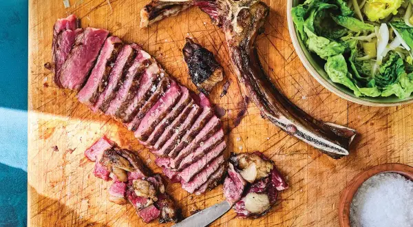 A perfectly cooked Spanish steak, sliced and ready to enjoy, sits on a wooden board with a side of fresh salad and a bowl of coarse salt.